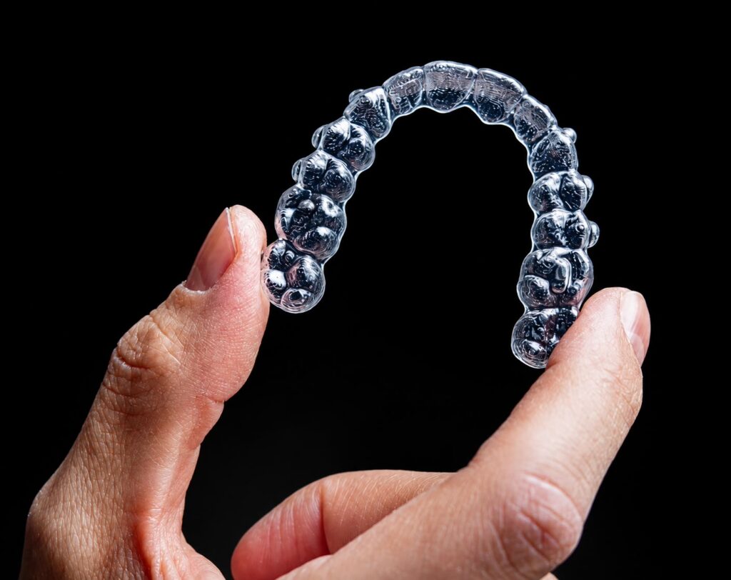 Transparent removable orthodontic aligner held between finger and thumb against a black background